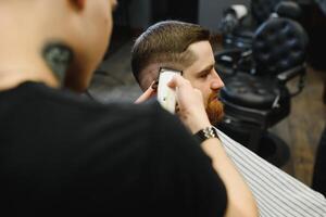 Herstellung Haarschnitt aussehen perfekt. jung bärtig Mann bekommen Haarschnitt durch Friseur während Sitzung im Stuhl beim Friseurladen. foto