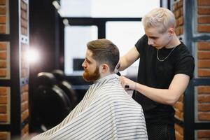 Herstellung Haarschnitt aussehen perfekt. jung bärtig Mann bekommen Haarschnitt durch Friseur während Sitzung im Stuhl beim Friseurladen. foto
