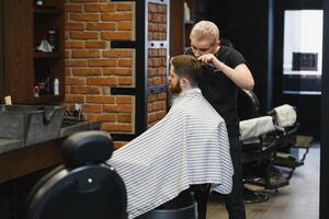 Herstellung Haarschnitt aussehen perfekt. jung bärtig Mann bekommen Haarschnitt durch Friseur während Sitzung im Stuhl beim Friseurladen. foto