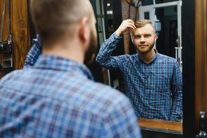 Perfektion. gut aussehend bärtig Mann beim Friseurladen. Porträt von stilvoll jung Kerl im Jacke berühren seine Haar während Besuch Friseur oder Salon. Herren Haarschnitt. Friseur Konzept. foto