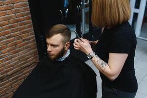 stilvoll Mann Sitzung Barbier Geschäft Friseur Friseur Frau Schneiden seine Haar Porträt gut aussehend glücklich jung bärtig kaukasisch Kerl bekommen modisch Haarschnitt attraktiv Barbier Mädchen Arbeiten Portion Klient foto