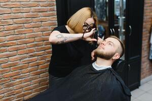 jung Erwachsene schön kaukasisch Frau Friseur schneidet Bart gut aussehend Mann beim modern Friseur glücklich Fröhlich Sitzung Stuhl Männer Schönheit Salon. Bart Pflege. metrosexuell. weiblich Barbier im schwarz Handschuhe Portion foto