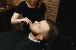 jung Erwachsene schön kaukasisch Frau Friseur schneidet Bart gut aussehend Mann beim modern Friseur glücklich Fröhlich Sitzung Stuhl Männer Schönheit Salon. Bart Pflege. metrosexuell. weiblich Barbier im schwarz Handschuhe Portion foto