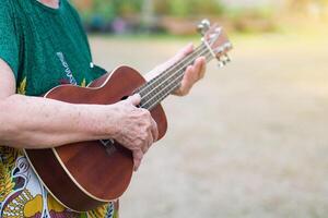 Nahansicht von Hände Senior Frau spielen das Ukulele während Stehen im ein Garten. entspannend durch abspielen klein Gitarre glücklich. selektiv Fokus. Raum zum Text. Konzept von alt Menschen und Entspannung foto