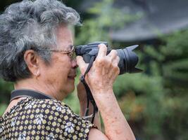 Senior Frau mit kurz grau Haar tragen Brille und Schießen Fotografie durch ein Digital Kamera beim das Park. Konzept von alt Menschen und Fotografie foto
