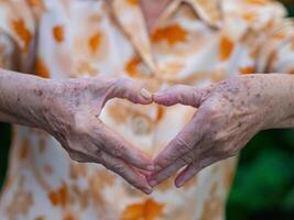 Nahansicht von Hand Senior Frau Herstellung ein Herz Symbol während Stehen im das Garten. Raum zum Text. Konzept von alt Menschen und Gesundheitswesen foto