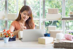 Geschäftsfrau lange Haar tragen ein beiläufig Stoff und schreiben Daten auf Startseite Paket Box zum bereiten senden zu das Kunde während Sitzung auf ein Stuhl beim das Büro. Konzept von Geschäft und E-Commerce foto