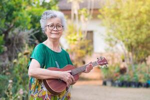 Porträt von ein Alten Frau spielen das Ukulele während Stehen im das Garten. entspannend durch Singen und abspielen klein Gitarre glücklich und genießen Leben nach in den Ruhestand gehen. Konzept von alt Menschen und Gesundheit Pflege foto