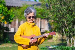 Porträt von ein Senior Frau mit kurz grau Haar spielen das Ukulele, lächelnd, und suchen beim das Kamera während Stehen im ein Garten. Konzept von alt Menschen und Entspannung foto