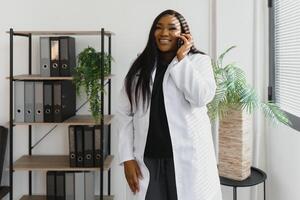 Medizin, Technologie und Gesundheitswesen Konzept - - glücklich lächelnd afrikanisch amerikanisch weiblich Arzt oder im Weiß Mantel mit Stethoskop Berufung auf Smartphone Über Krankenhaus Hintergrund. foto