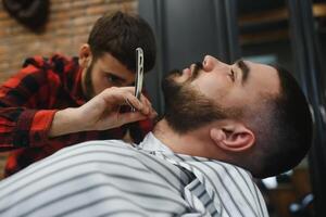 ernst bärtig Mann bekommen Bart Haarschnitt mit ein Gerade Rasierer durch Barbier während Sitzung im Stuhl beim Friseurladen. Friseur Thema. foto