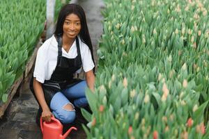 schön jung lächelnd afrikanisch amerikanisch Mädchen, Arbeiter mit Blumen im Gewächshaus. Konzept Arbeit im das Gewächshaus, Blumen. foto