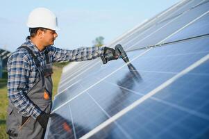 Porträt von jung indisch Mann Techniker tragen Weiß schwer Hut Stehen in der Nähe von Solar- Paneele gegen Blau Himmel. industriell Arbeiter Solar- System Installation foto