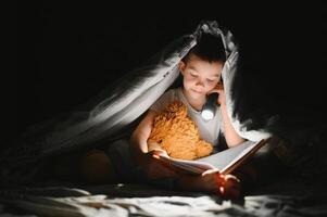 Kind lesen Buch im Bett. Kinder lesen beim Nacht. wenig Junge mit Fee Geschichte Bücher im Schlafzimmer . Bildung zum jung Kinder. Bettzeit Geschichte im das Abend. süß Kind unter Decke im dunkel Zimmer mit Lampe foto