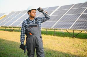 Porträt von jung indisch Mann Techniker tragen Weiß schwer Hut Stehen in der Nähe von Solar- Paneele gegen Blau Himmel. industriell Arbeiter Solar- System Installation foto