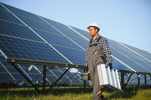 Porträt von jung indisch Mann Techniker tragen Weiß schwer Hut Stehen in der Nähe von Solar- Paneele gegen Blau Himmel. industriell Arbeiter Solar- System Installation foto