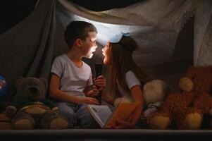 zwei wenig Kind abspielen beim Zuhause im das Abend zu bauen ein Camping Zelt zu lesen Bücher mit ein Taschenlampe und Schlaf innen. foto