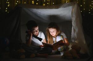 zwei Kinder mit Taschenlampe lesen ein Buch unter ein Decke wie ein Zelt foto