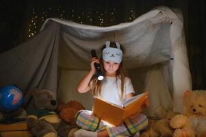 Kind Mädchen lesen mit Buch und Taschenlampe und Teddy Bär im Zelt. Vor gehen zu Bett foto