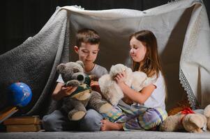 zwei wenig Kinder abspielen beim Zuhause im das Abend zu bauen ein Camping Zelt zu lesen Bücher mit ein Taschenlampe und Schlaf Innerhalb foto