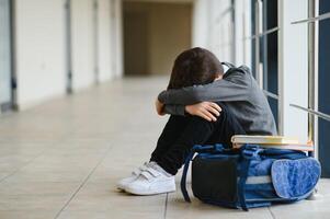 verärgert Junge Sitzung beim Schule und Weinen nach Tyrannisieren durch Schüler Klassenkameraden. das Kind bedeckt seine Gesicht mit seine Hände und weint foto