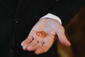 das Bräutigam auf seine Hochzeit Tag hält zwei Hochzeit Ringe im seine Hand im ein Hotel Zimmer. das Mann ist tragen ein Weiß Hemd und Blau Hose mit ein Weste. männlich Hand und Hochzeit Ringe schließen hoch. foto