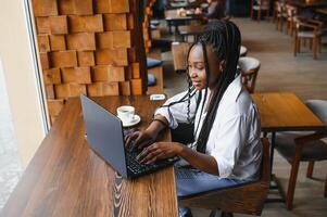 schön afrikanisch Geschäft Frau warten zum das Treffen im ein zusammenarbeiten Raum und Sitzung mit das tragbar Computer. Hipster Mädchen ist Surfen das Netz auf ein Laptop während Mittagessen. foto