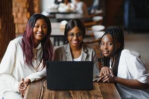 drei afrikanisch amerikanisch Mädchen Sitzung beim das Tabelle im Cafe studieren oben zum Prüfung oder Herstellung Hausaufgaben zusammen, Sie sind mit Laptop und Digital Tablette. foto