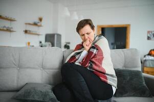 mittleren Alters 50er Jahre krank gefroren Frau sitzend auf Sofa im Leben Zimmer bedeckt mit warm Plaid Niesen, flüssig Nase fühlt sich ungesund, saisonal kalt, geschwächt immun System Konzept. foto