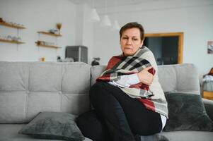 mittleren Alters 50er Jahre krank gefroren Frau sitzend auf Sofa im Leben Zimmer bedeckt mit warm Plaid Niesen, flüssig Nase fühlt sich ungesund, saisonal kalt, geschwächt immun System Konzept. foto