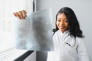 jung attraktiv afrikanisch weiblich Arzt Radiologe suchen beim Röntgen Bild, während Stehen im Licht modern Büro im Klinik foto