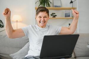 aufgeregt reifen Frau suchen beim Laptop Bildschirm, lesen gut Nachrichten im Nachricht, feiern online Lotterie gewinnen, Jubel Erfolg, überglücklich älter weiblich Sitzung auf Couch beim heim, mit Computer. foto