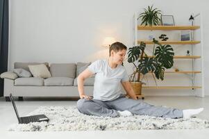 Fitness beim Zuhause online. Mitte alt Frau im Sportbekleidung tun Yoga auf Sport Matte mit Laptop im Leben Zimmer Innere mit Geräte, Profil, kostenlos Raum. foto