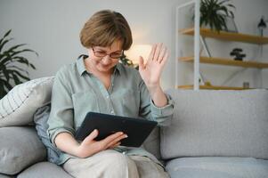 Pensionierung Freizeit. lächelnd Alten Frau ruhen mit Digital Tablette auf Couch beim heim, Surfen Internet oder Aufpassen Filme foto