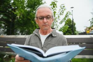 alt Mann mit grau Haar liest ein Buch auf ein Bank im das Park. sich ausruhen im das Park. foto