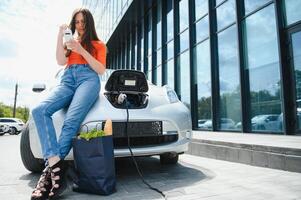 mit Smartphone während warten. Frau auf das elektrisch Autos aufladen Bahnhof beim Tageszeit. Marke Neu Fahrzeug. foto