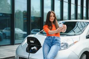 mit Smartphone während warten. Frau auf das elektrisch Autos aufladen Bahnhof beim Tageszeit. Marke Neu Fahrzeug. foto