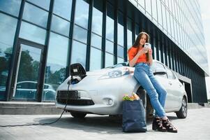 mit Smartphone während warten. Frau auf das elektrisch Autos aufladen Bahnhof beim Tageszeit. Marke Neu Fahrzeug. foto