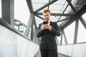 glücklich lächelnd Geschäftsmann tragen schwarz passen und mit modern Smartphone in der Nähe von Büro beim früh Morgen, erfolgreich Arbeitgeber zu machen ein Deal während Stehen in der Nähe von Wolkenkratzer Büro beim Nacht, Fackel Licht foto