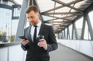 bärtig Geschäftsmann im formal passen auf brechen mit Handy, Mobiltelefon Telefon verwenden Smartphone. Geschäft Mann Stehen draußen auf modern städtisch Stadt Straße Hintergrund mit Kaffee Tasse im Innenstadt draußen. Kopieren Raum foto