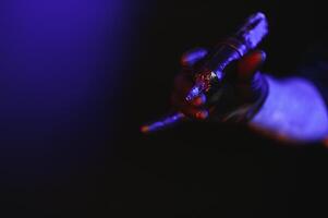 Nahansicht von Tätowierer Meister Hand im schwarz Handschuh halten Maschine zum Herstellung tätowieren Kunst auf Körper isoliert auf dunkel Hintergrund im Neon. foto