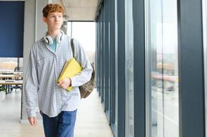 Porträt von lächelnd Clever lockig behaart Teenager Junge halten Buch suchen beim Kamera. zurück zu Schule, Bildung Konzept foto
