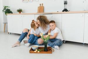 glücklich Mutter umarmen Tochter und Sohn Sitzung auf hölzern Fußboden im modern Küche beim heim. Mama umarmen Kinder. glücklich Familie Beziehung Konzept foto