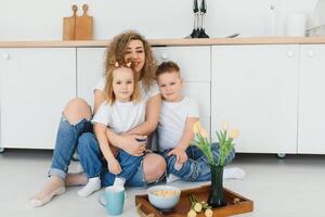 glücklich Mutter umarmen Tochter und Sohn Sitzung auf hölzern Fußboden im modern Küche beim heim. Mama umarmen Kinder. glücklich Familie Beziehung Konzept foto