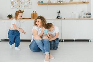 glücklich jung Familie, Mama mit wenig Kinder sitzen auf warm hölzern Fußboden im Neu modern Design Küche, Mutter mit aufgeregt klein Kinder entspannen sich ausruhen im besitzen renoviert Wohnung, ziehen um Konzept foto