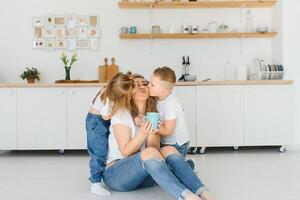 glücklich jung Familie, Mama mit wenig Kinder sitzen auf warm hölzern Fußboden im Neu modern Design Küche, Mutter mit aufgeregt klein Kinder entspannen sich ausruhen im besitzen renoviert Wohnung, ziehen um Konzept foto