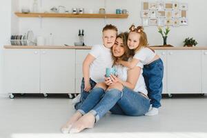 glücklich jung Familie, Mama mit wenig Kinder sitzen auf warm hölzern Fußboden im Neu modern Design Küche, Mutter mit aufgeregt klein Kinder entspannen sich ausruhen im besitzen renoviert Wohnung, ziehen um Konzept foto