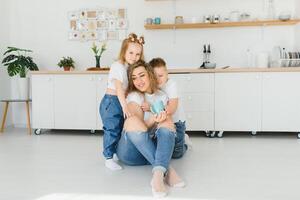 glücklich jung Familie, Mama mit wenig Kinder sitzen auf warm hölzern Fußboden im Neu modern Design Küche, Mutter mit aufgeregt klein Kinder entspannen sich ausruhen im besitzen renoviert Wohnung, ziehen um Konzept foto