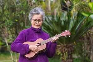 Porträt von ein glücklich Senior Frau spielen das Ukulele, lächelnd und suchen Nieder während Stehen im ein Garten. Raum zum Text. Konzept von alt Menschen und Entspannung foto