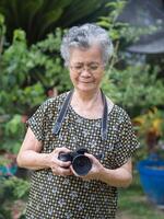 Senior Frau mit kurz grau Haar tragen Brille und Schießen Fotografie durch ein Digital Kamera beim das Garten. Konzept von alt Menschen und Fotografie foto